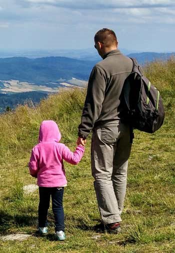 A father and his daughter hold hands and look down from a mountain. 