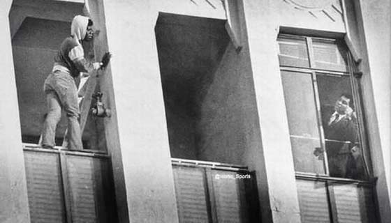 Mohammad Ali standing at a window talking to a man standing on a window sill a few feet away.