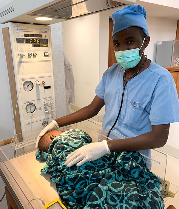 man in blue doctors scrubs caring for a baby wrapped in blue and black African fabric