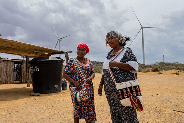 Two wayuu women stand with wind mills in the background