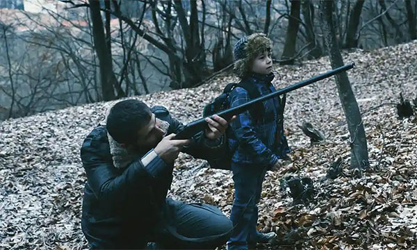 Still from the film RMN. A father aims a gun in the gloomy woods with his young son at his side. 