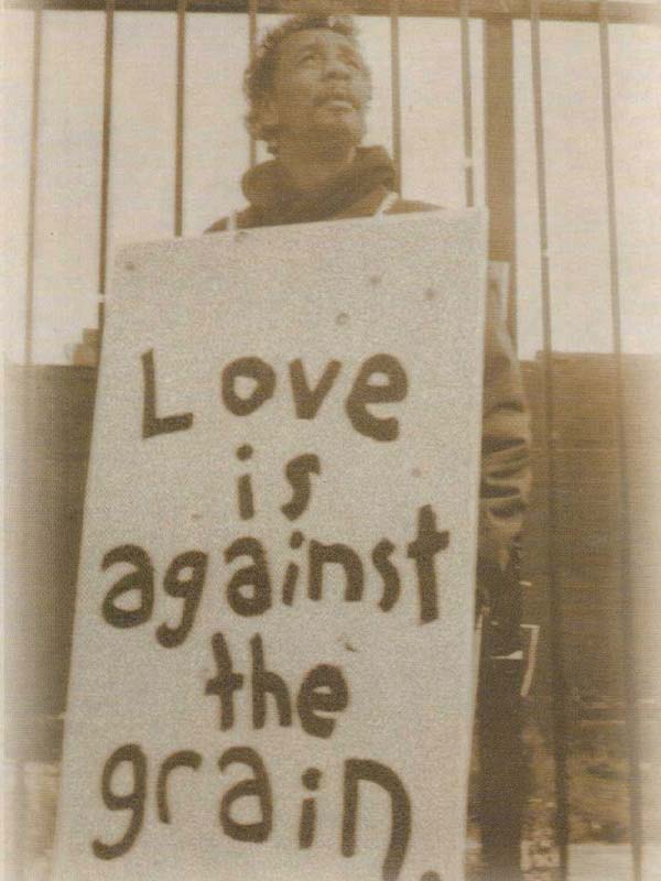 Man leaning against bars with a sandwich board reading "Love is against the grain."