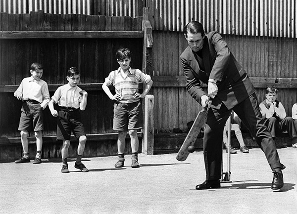 A cricket player swings a bat while young boys look on
