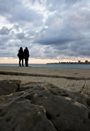 two people standing on a rocky beach looking at a city