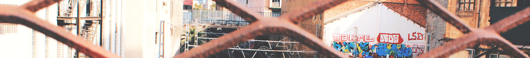 Part of a photograph by Juul Evers showing buildings, train tracks, and graffiti through a metal fence.