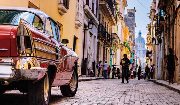 A red, old-fashioned car on a cobblestoned Havana street lined with colorful houses; the capitol building visible through the gap the end of the street.