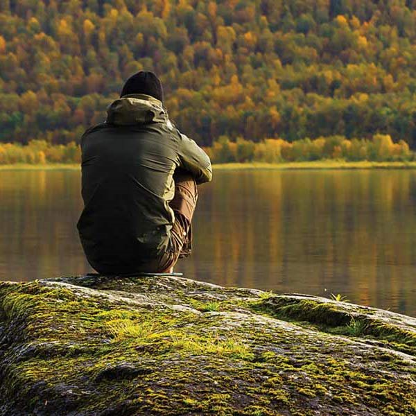 Man sitting on a rock looking at reflection of autumn trees in lake