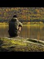 Man sitting on a rock looking at reflection of autumn trees in lake