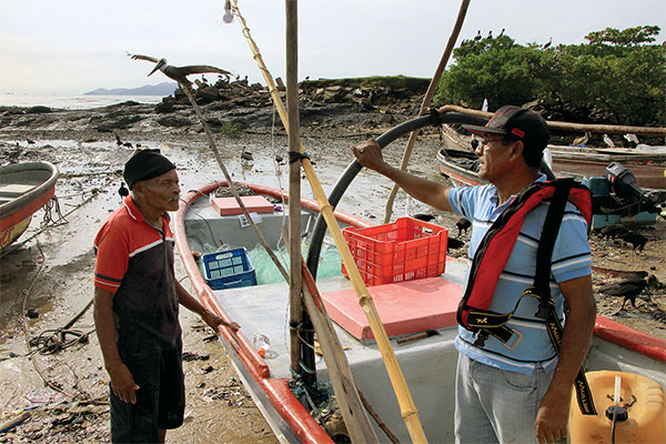 two older men talking on a beach while leaning on a small boat