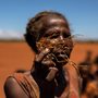 a woman holds part of a dead corn plant in a field covered with red sand